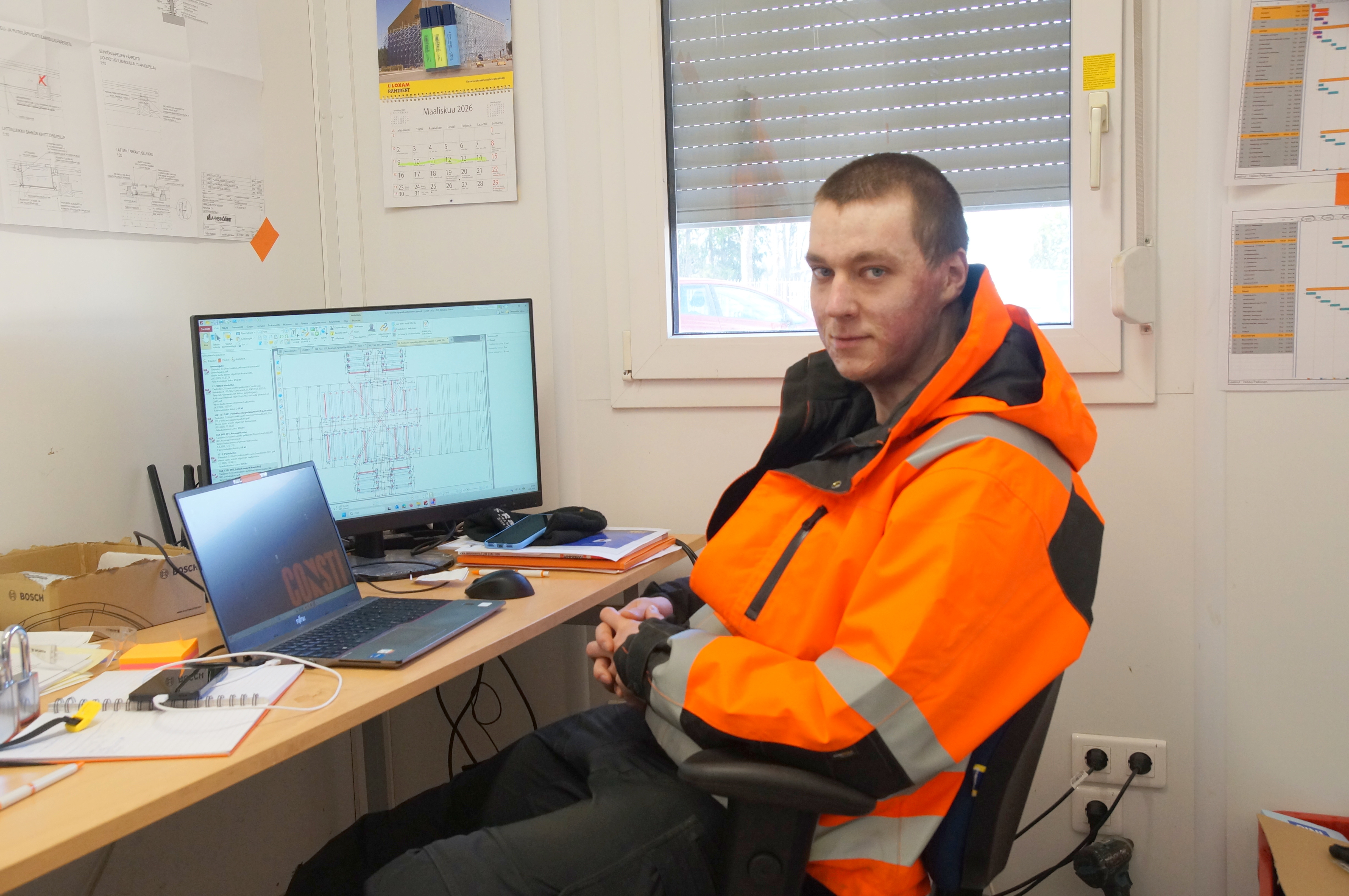 Oranssitakkinen mies työpöydän ääressä. Pöydällä on kannettava tietokone ja erillinen näyttö.