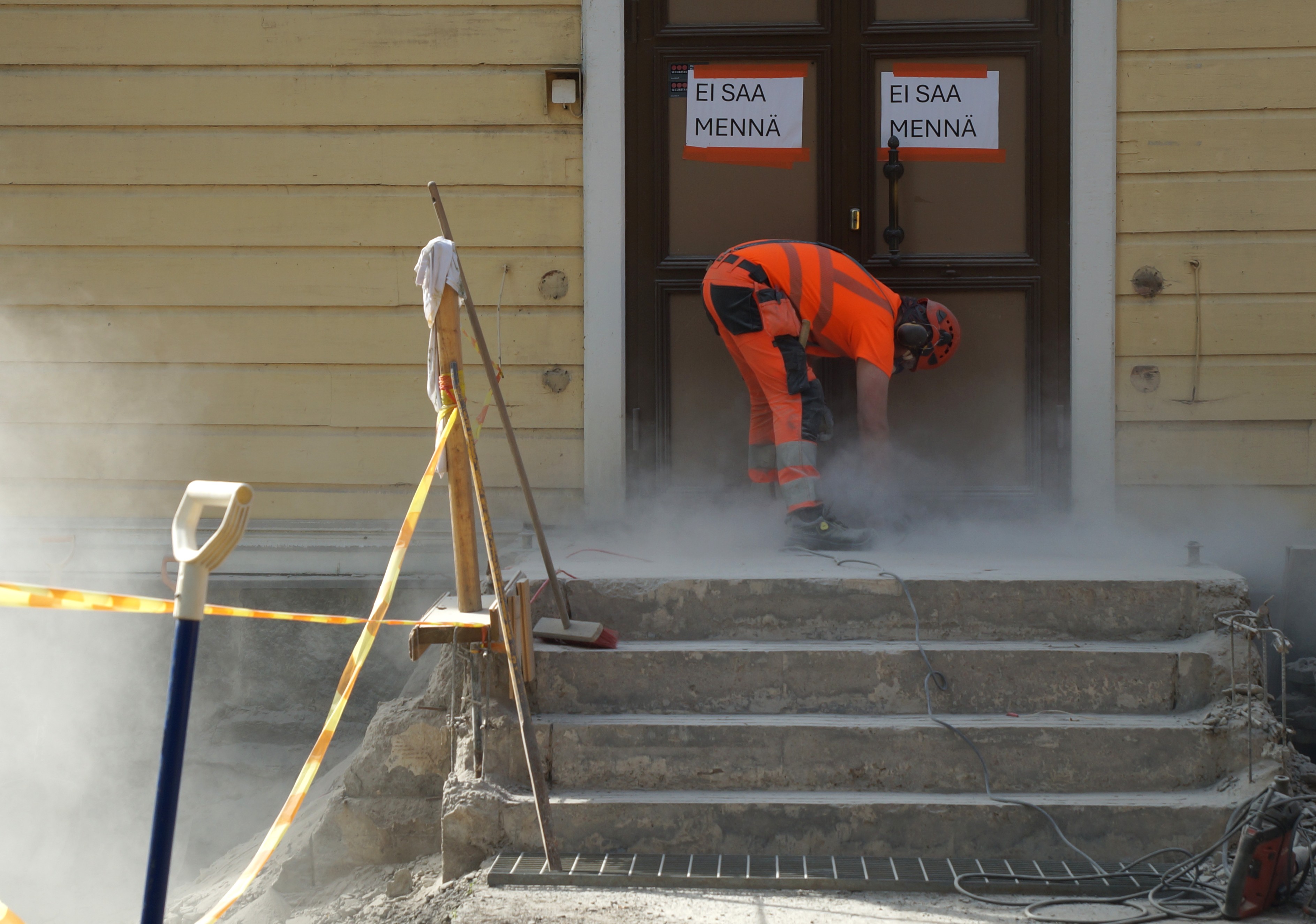 Oranssiasuinen työmies hioo kirkon portaita ja pölyävä laasti näkyy kirkon seinää vasten.