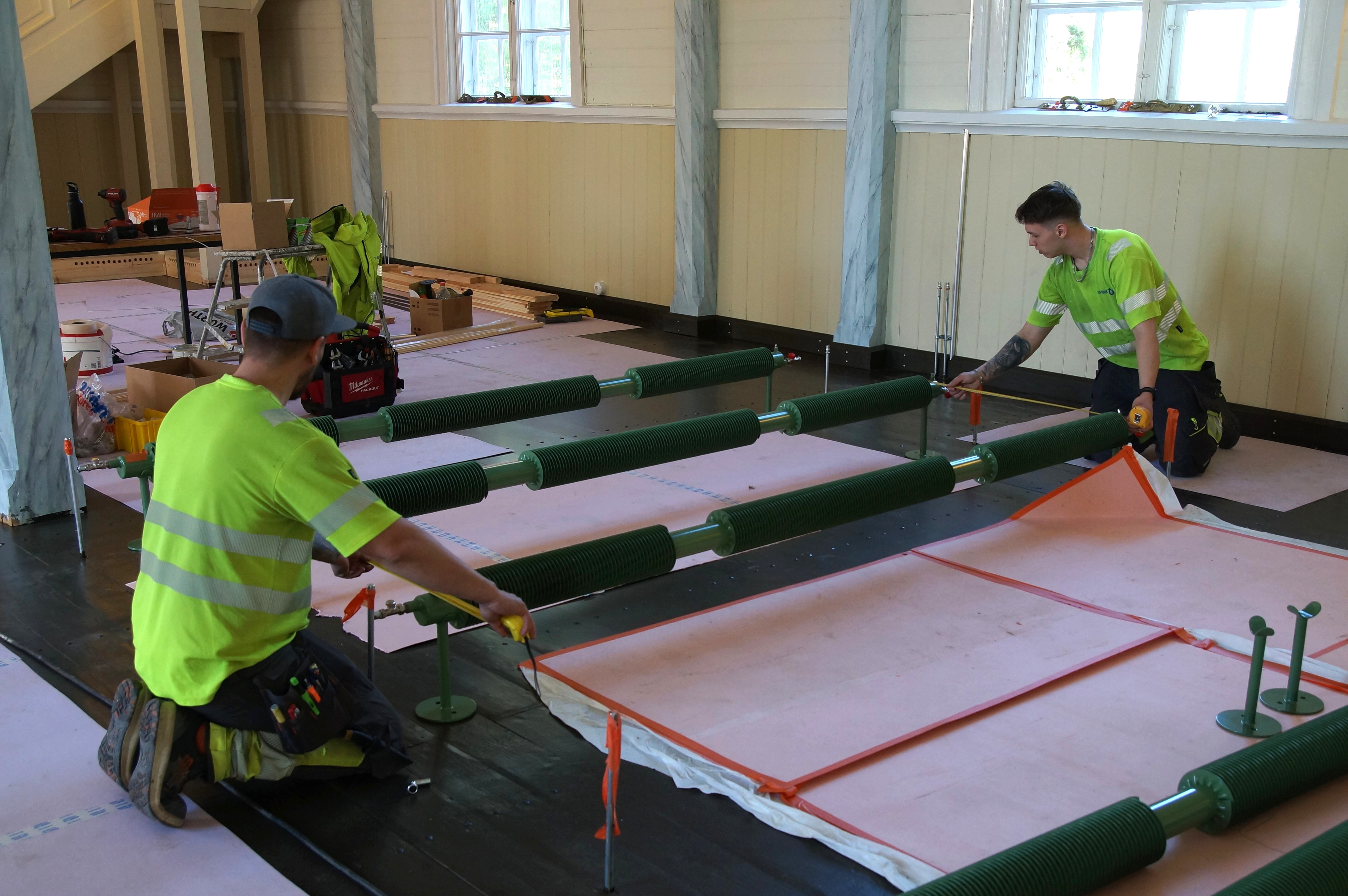 Kuvassa näkyy kaksi työmiestä, jotka työskentelevät polvillaan kirkon lattialla. Heidän välissään on pitkä vihreä lämpöpatteri, josta he mittaavat etäisyyttä seuraavaan patteriin.