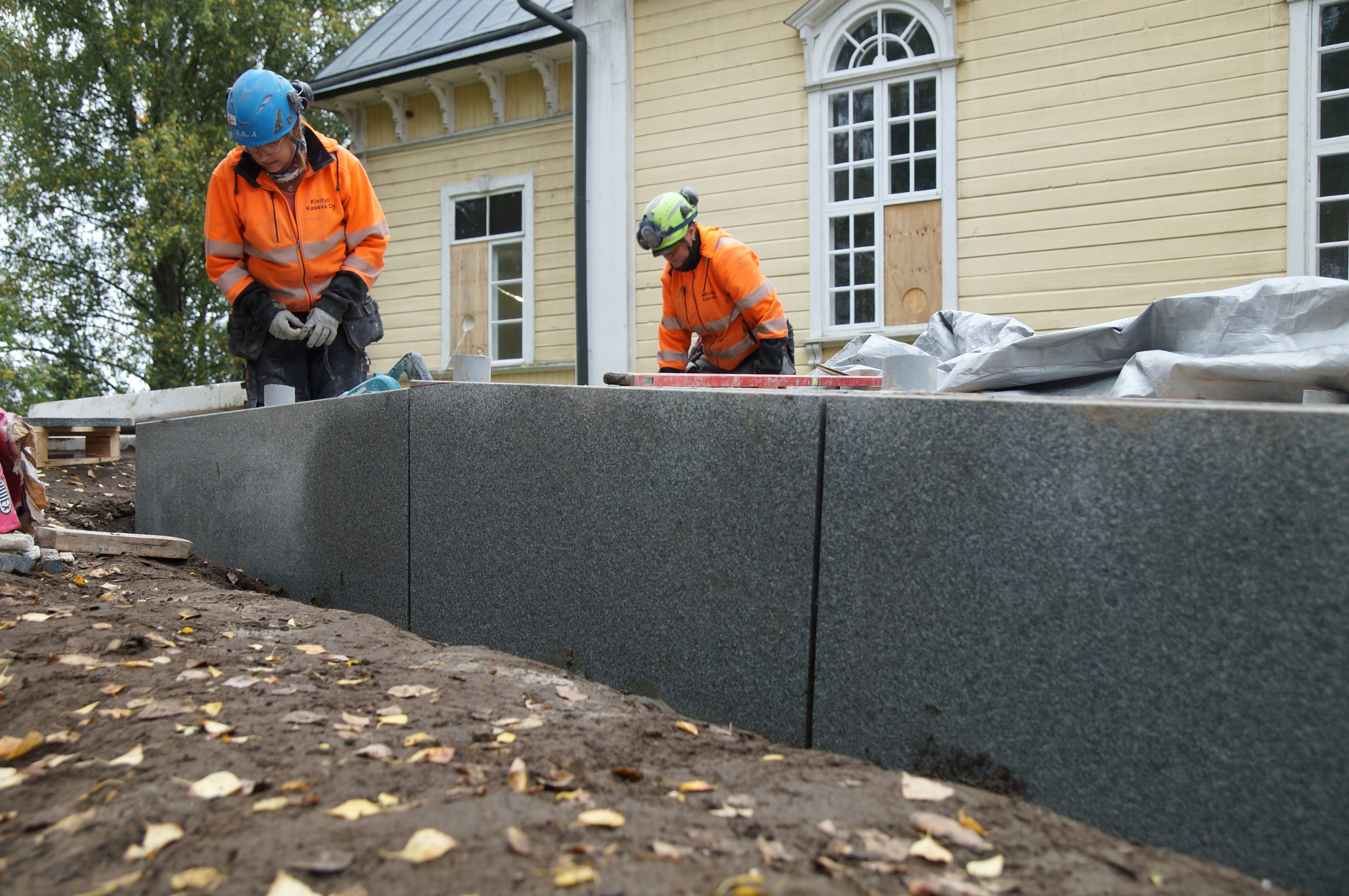 Kuvassa näkyy kolme harmaata graniittilevyä, joiden takan takana on kirkon invaluiska. Kaksi työntekijää katsoo luiskan päällä, miltä työ näyttää.