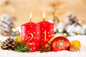 Kaksi adventtikynttilää palavat joulukoristeiden keskellä. Kynttilöissä on numerot 1 ja 2 ja ne ovat punaisia.