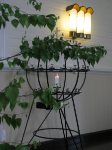 Kirkon lähetyskynttelikkö, jossa palaa yksi kynttilä, edessä koivunoksia.