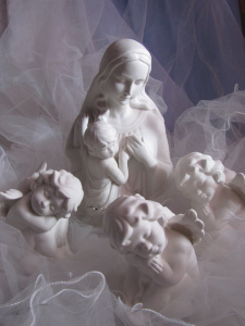 Posliininen Maria-patsas Jeesus-lapsi sylissään sekä kolme posliinista enkeliä. Taustalla vaaleansinistä pitsiä.
