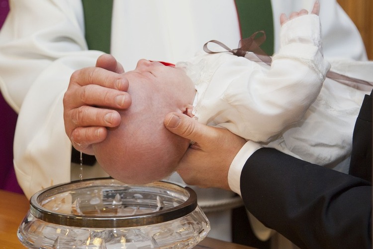 Pappi kastaa vauvaa kastelemalla vettä tämän päähän . Kaste liittää meidät Jumalan perheeseen.