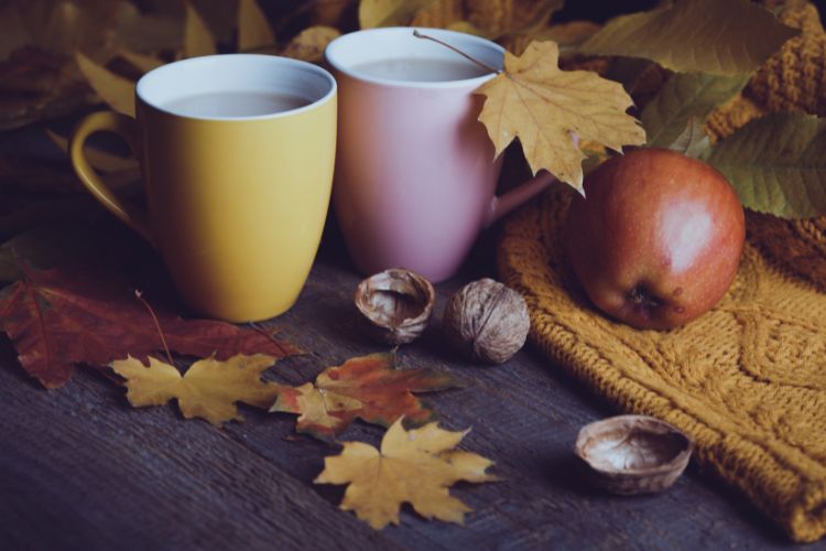 Kaksi kahvikuppia, syksyisiä lehtiä, pähkinänkuoria, omena ja neule pöydällä.